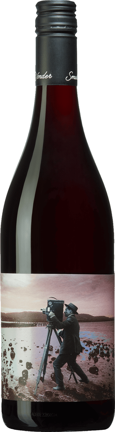 Flaska med mörkt glas, svart kapsyl och en etikett med ett gammalt foto av en man med kamera på stativ vid vattnet och texten Small Wonder. Small Wonder Landscape Pinot Noir, flaska. Flaska med mörkt glas, svart kapsyl och en etikett med ett gammalt foto av en man med kamera på stativ vid vattnet och texten Small Wonder.